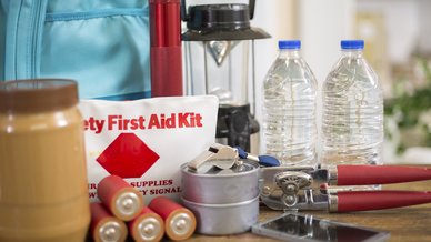 An emergency preparedness kit with a first aid bag, water bottles, batteries, a lantern, can opener and backpack on a table.