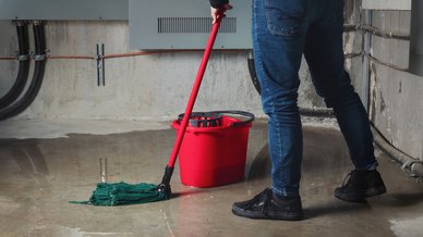 A person mopping standing water off a flooded basement floor with a red bucket nearby.